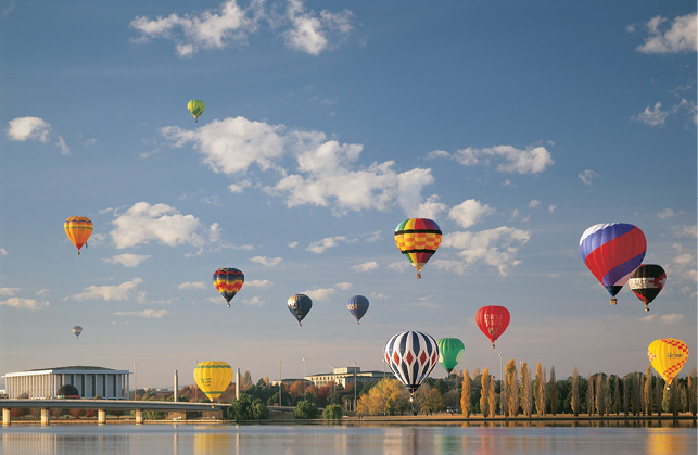 A number of colourful hot-air balloons are in the sky over the shores of a still lake. Significant buildings including the National Library of Australia are visible on the lake front.