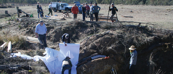 A group of people are standing at the edge of a paddock watching four men installing white fabric and logs into the steep bank of a gully below the paddock. There are two vehicles and piles of dressed pine logs in the paddock.