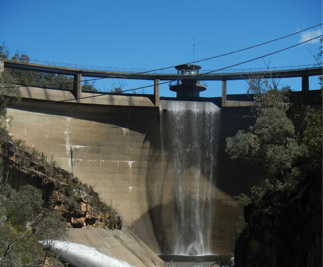 A curved concrete dam wall spans a narrow valley of rock. A small amount of water is spilling over a section of the dam.